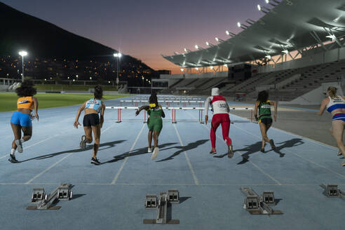 Female track and field athletes racing to hurdles on track at night - CAIF31793