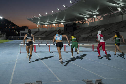 Female track and field athletes racing toward hurdles in stadium - CAIF31797