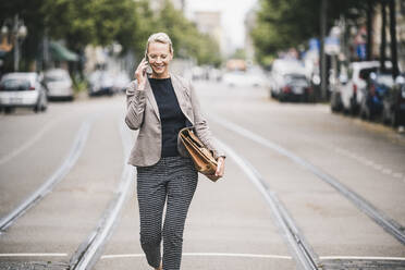 Smiling businesswoman holding bag while talking on mobile phone - UUF23907
