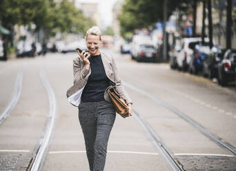 Cheerful female commuter talking through mobile phone while walking on tramway - UUF23909