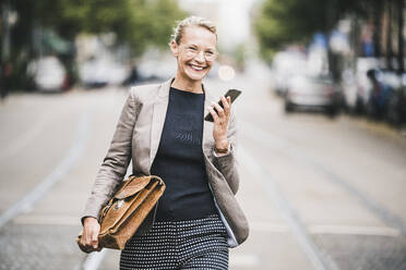 Cheerful businesswoman with smart phone and bag walking on street - UUF23910