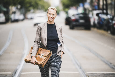 Lächelnde Geschäftsfrau mit Hand in der Tasche, die eine Tasche hält, während sie auf der Straße geht - UUF23912
