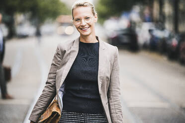 Lächelnde reife Geschäftsfrau, die eine Tasche hält, während sie auf der Straße steht - UUF23914