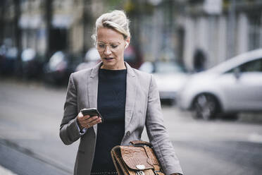 Blonde Pendlerin hält Tasche und benutzt Smartphone - UUF23921