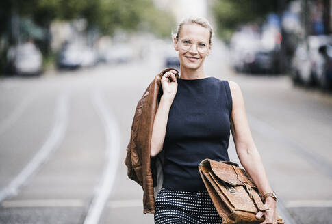 Lächelnde Geschäftsfrau mit Jacke und Tasche beim Gehen auf der Straße - UUF23924