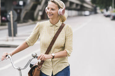 Blonde Geschäftsfrau mit Kopfhörern lächelt, während sie auf der Straße Fahrrad fährt - UUF23930