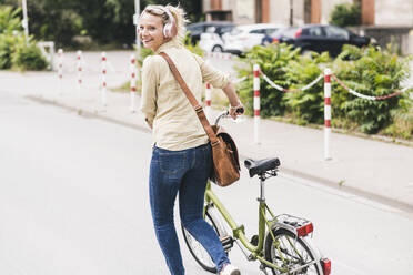 Happy female commuter wheeling bicycle while looking back over shoulder - UUF23931