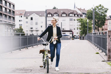 Mature businesswoman wheeling bicycle on bridge in city - UUF23933