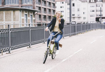 Glückliche Pendlerin, die mit ihrem Handy spricht, während sie auf einer Brücke Fahrrad fährt - UUF23944