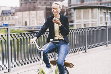 Fröhliche Geschäftsfrau, die über ihr Handy spricht, während sie auf einer Brücke Fahrrad fährt - UUF23945