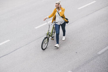Female commuter listening music through headphones while wheeling bicycle on street - UUF23957