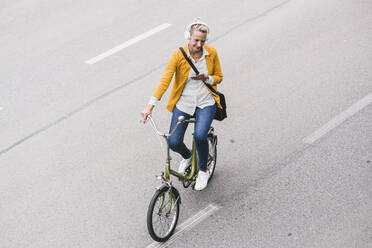 Ältere Geschäftsfrau mit Kopfhörern und Mobiltelefon fährt Fahrrad auf der Straße - UUF23958