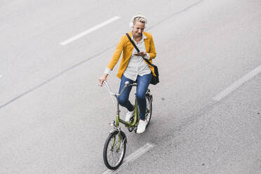 Mature businesswoman with headphones and mobile phone riding bicycle on street - UUF23958