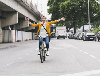 Carefree female commuter with arms outstretched riding bicycle on street - UUF23960