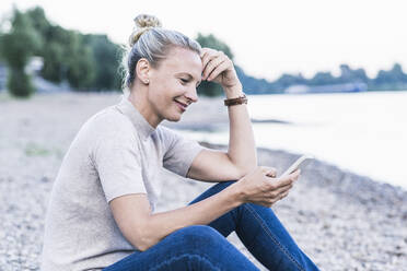 Woman using mobile phone while sitting at riverbank - UUF23975