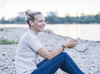 Happy mature woman holding mobile phone while sitting at riverbank - UUF23977