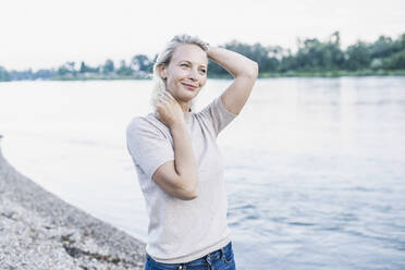Blonde Frau mit Hand im Haar steht am Flussufer - UUF23979