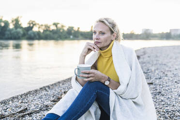 Thoughtful mature woman holding coffee cup while sitting by riverbank - UUF23986