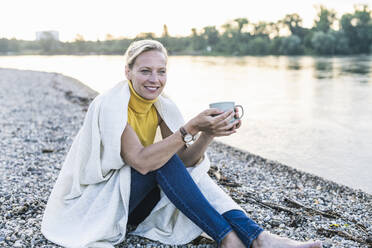 Ältere Frau mit Kaffeetasse am Flussufer sitzend - UUF23987