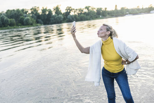 Frau knutscht, während sie ein Selfie mit ihrem Smartphone am Flussufer macht - UUF23994