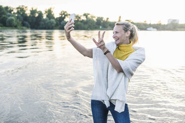Smiling woman gesturing peace sign while taking selfie through smart phone at riverbank - UUF23996