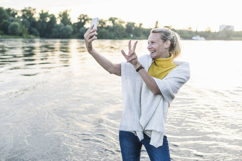 Lächelnde Frau gestikuliert Friedenszeichen bei der Aufnahme von Selfie durch Smartphone am Flussufer - UUF23996