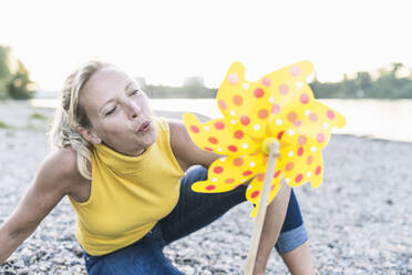 Playful woman blowing pinwheel toy while sitting at riverbank - UUF23998