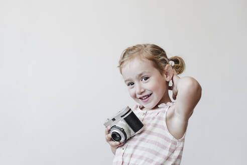 Lächelndes blondes Mädchen hält Spielzeugkamera vor weißem Hintergrund - EBBF04194