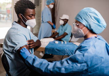 Seitenansicht einer Ärztin in Schutzkleidung und Latexhandschuhen, die ihren Arm mit alkoholhaltiger Baumwolle desinfiziert, um einen männlichen afroamerikanischen Patienten in einer Klinik während des Ausbruchs des Coronavirus zu impfen - ADSF25591
