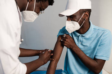 Schwarzer Arzt in Schutzkleidung und Latexhandschuhen bei der Impfung eines männlichen afroamerikanischen Patienten in einer Klinik während des Ausbruchs des Coronavirus - ADSF25599
