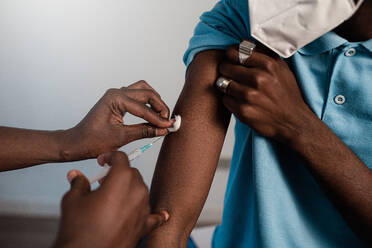 Unkenntlich gemachter schwarzer Facharzt desinfiziert Arm mit Watte mit Alkohol, um männlichen afroamerikanischen Patienten in der Klinik während des Ausbruchs des Coronavirus zu impfen - ADSF25600