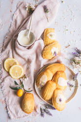 Draufsicht auf leckere Madeleines auf einem Teller zwischen frischen Zitronenscheiben und blühenden Lavendelzweigen auf zerknittertem Textil - ADSF25615