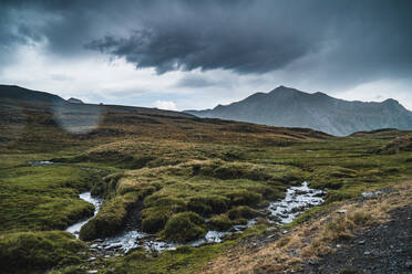 Breathtaking landscape of meadow with green grass and creek flowing on hills under gloomy sky in Pyrenees in Spain - ADSF25634