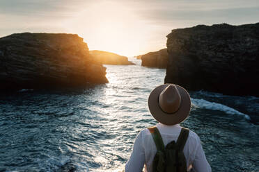 Man with backpack and hat standing and looking at the sea - ADSF25649