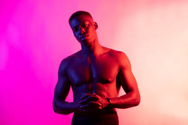 Ernster junger afroamerikanischer männlicher Sportler mit nacktem Oberkörper, der mit gefalteten Händen auf rosa Hintergrund in einem Neonstudio in die Kamera schaut - ADSF25652