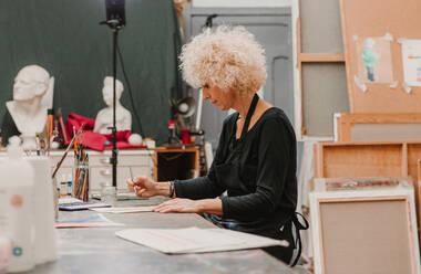 Konzentrierte Künstlerin sitzt am Tisch und malt mit Aquarellfarben auf Papier, während sie in einer Kreativwerkstatt arbeitet - ADSF25657