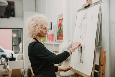 Weibliche Künstlerin zeichnet einen Menschen mit Bleistift, während sie an einer Staffelei im Atelier steht - ADSF25665