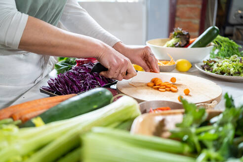 Eine nicht erkennbare Frau schneidet eine rohe Karotte mit einem Messer, während sie ein vegetarisches Essen im Haus zubereitet - ADSF25672
