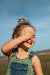 Delighted adorable little girl in overalls standing with hand on face in meadow and closed eyes - ADSF25675