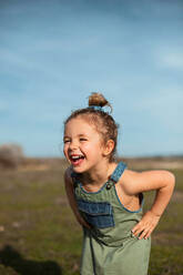 Delighted adorable little girl in overalls standing with hands on waist in meadow and looking away - ADSF25676