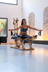 Konzentrierte, fitte Frau, die ihre Beine streckt und Ausfallschritte auf dem Pilates-Reformer während des Trainings im Fitnessstudio macht - ADSF25682