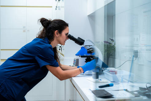 Side view of female vet in blue uniform looking through microscope while working in lab - ADSF25686