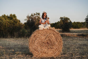 Woman contemplating while sitting with dog on straw bale during sunset - EBBF04206