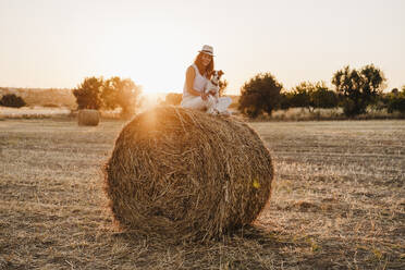 Smiling woman with Jack Russell Terrier sitting cross-legged on straw bale during sunset - EBBF04208