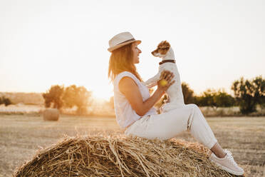 Woman wearing hat playing with dog on strawb bale - EBBF04209