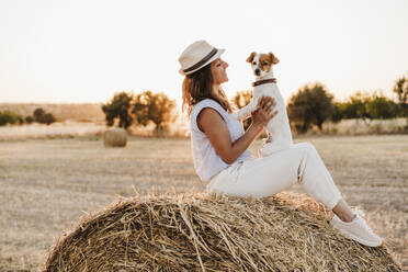 Smiling woman looking at dog while sitting on straw bale during sunset - EBBF04210