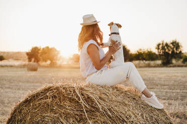 Woman with dog sitting on straw bale at farm during sunset - EBBF04211