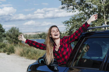 Cheerful young woman with arms outstretched leaning from car window enjoying sunny day - EBBF04216