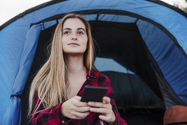 Nachdenkliche blonde Frau mit Smartphone vor blauem Zelt - EBBF04218