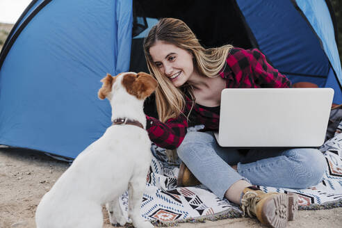 Glücklicher junger digitaler Nomade, der seinen Hund streichelt, während er mit seinem Laptop vor dem Zelt sitzt - EBBF04219